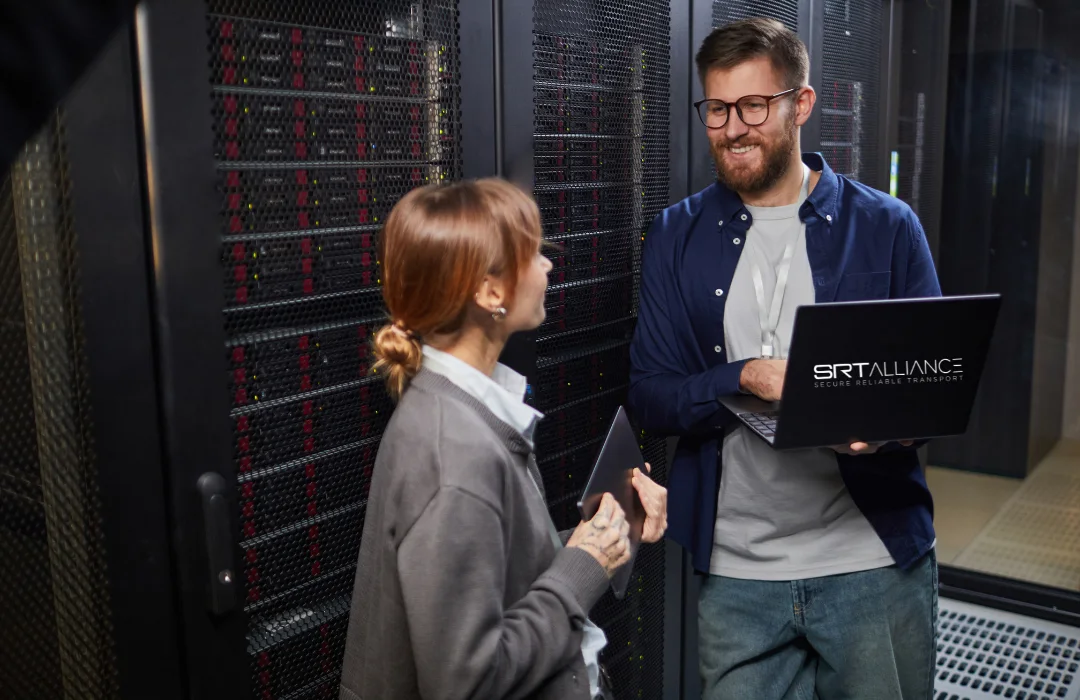 Two engineers talking beside server racks in a data centre, one holding an open laptop.